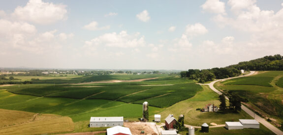 Farm buildings seen from above, encircled by green fields beneath a sky with scattered clouds.