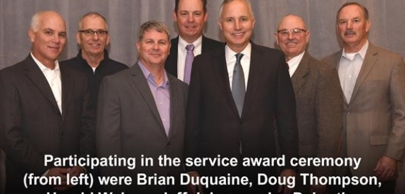 Seven men in suits stand side by side at a service award ceremony before a gray curtain.