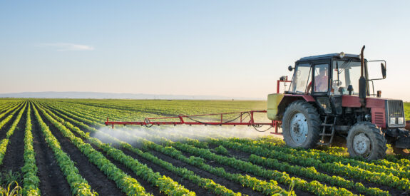 Red tractor spraying crops in a green field beneath a clear blue sky at sunset.