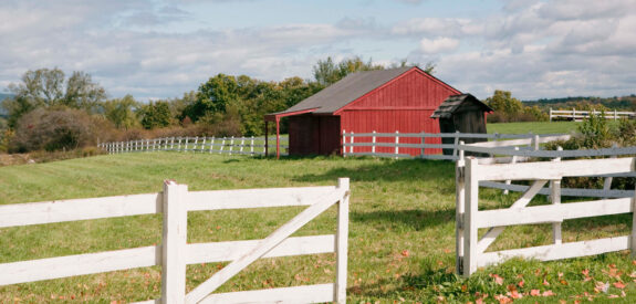 Red barn behind white fence on grassy field with autumn leaves under a partly cloudy sky.