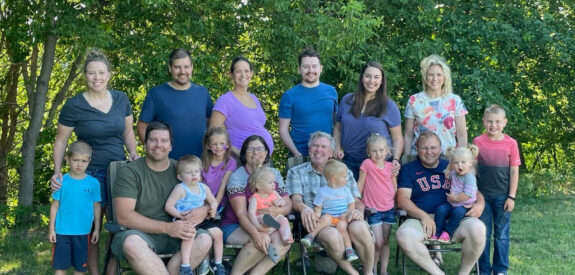 A big family with adults and kids poses outdoors in front of trees on a sunny day.