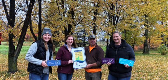 Four people outside in autumn hold signs promoting soil health, water quality, and climate change awareness.