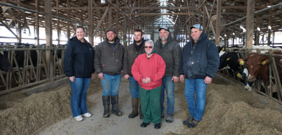 Six people pose in a barn, hay on the ground and cows lined up along both sides.