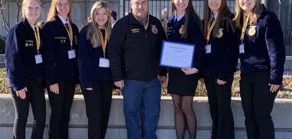 Seven people in FFA jackets stand outside, smiling; one person holds a certificate.