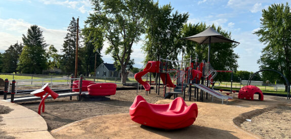 Playground featuring red climbing frames, slides, and benches among trees on a bright, sunny day.