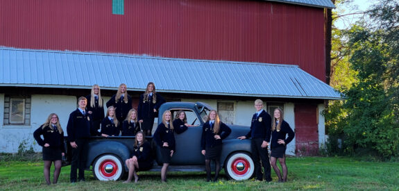 Twelve people in matching outfits pose on and around a vintage truck parked by a red barn.