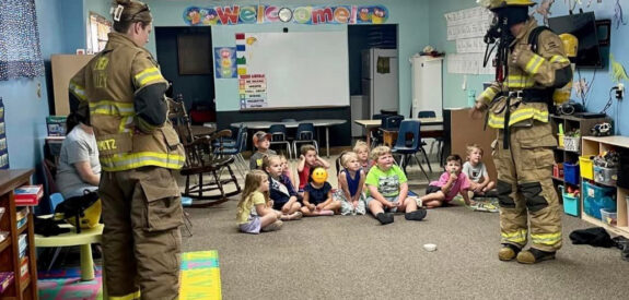 Firefighters in uniform speak to young children seated on a classroom floor.