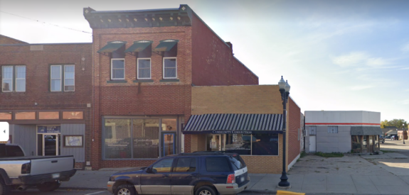 Red brick building with striped awning and large windows; several cars parked in front on a sunny day.