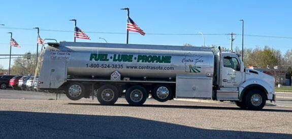Silver fuel delivery truck parked by a road with U.S. flags in the background on a sunny day.