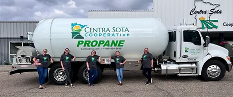 Five women in matching shirts stand beside a Centra Sota Cooperative propane truck outside a building.