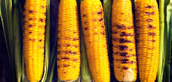 Grilled corn cobs with char marks rest on green husks atop a wooden table.