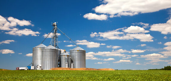 Silver grain silos stand in green crop fields beneath a blue sky with scattered clouds.