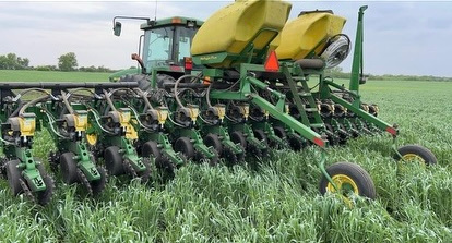 Green tractor with yellow planters planting seeds in a green field beneath a cloudy sky.