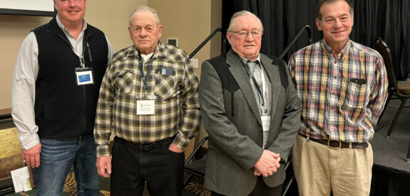 Four men indoors with conference badges; three wear plaid shirts, one wears a blazer.