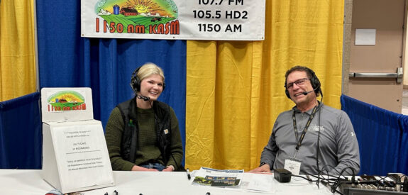 Two people with headsets smile in a radio station booth, celebrating 70 years of broadcasting.