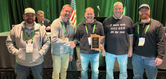 Five men indoors, one holding a plaque, with an American flag and green curtains behind them.