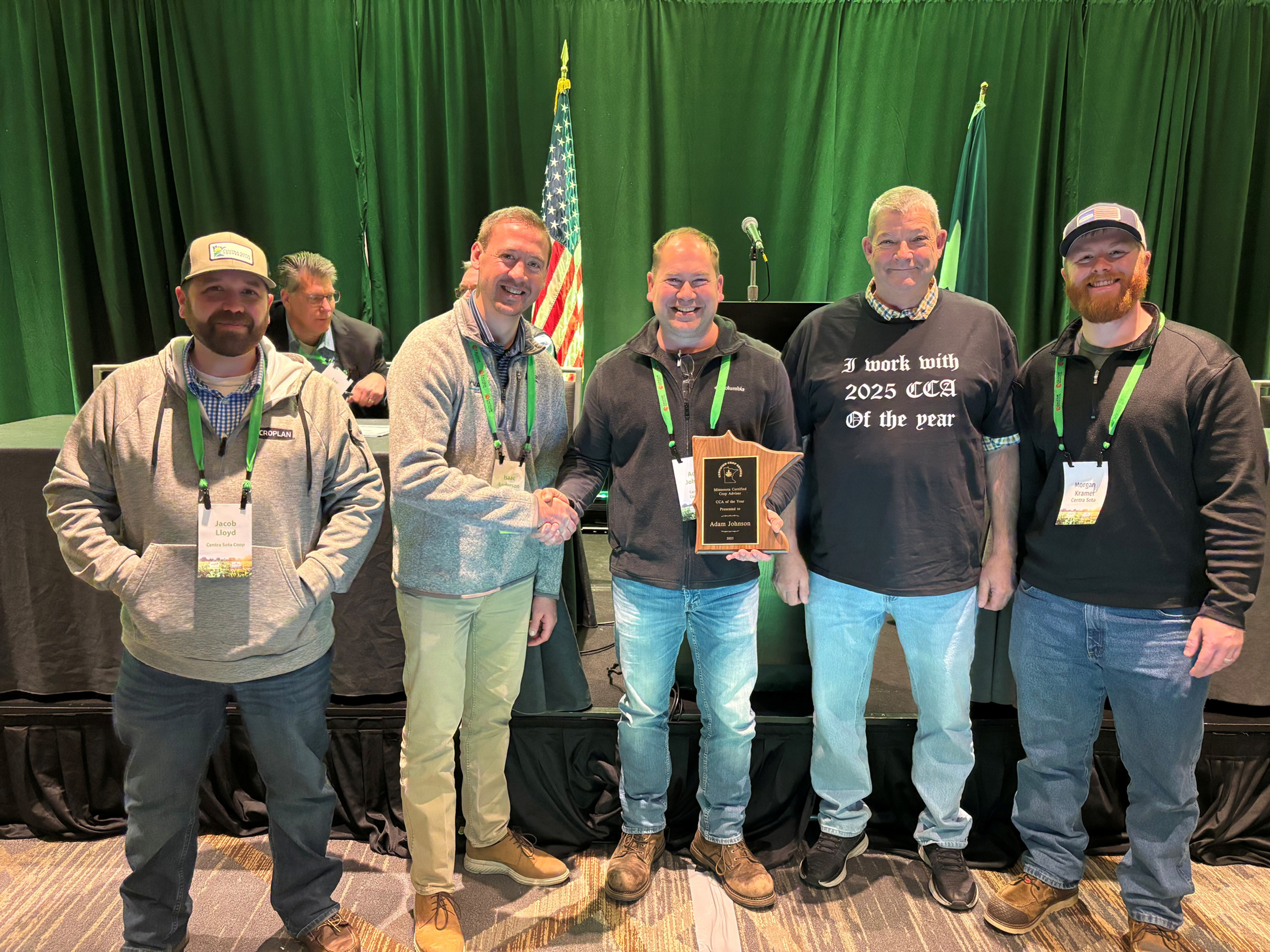 Five men smiling, one holding an award plaque. Green backdrop with an American flag. One shirt reads "I work with 2025 CCA of the year."