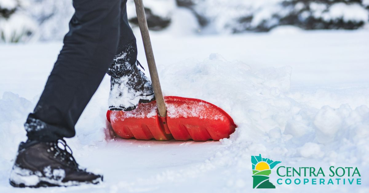 Person shoveling snow with a red shovel, wearing black pants and boots. Snowy background. Logo: Centra Sota Cooperative in corner.