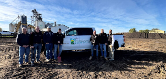 Eight people stand beside a Centra Sota Cooperative truck on a dirt lot, with buildings and equipment in the background.