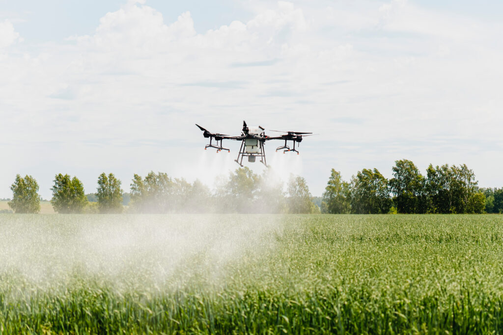 A drone sprays mist over green crops in a field, with trees and a cloudy sky behind.