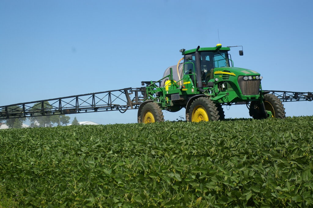A green sprayer tractor drives through a lush field beneath a clear blue sky.