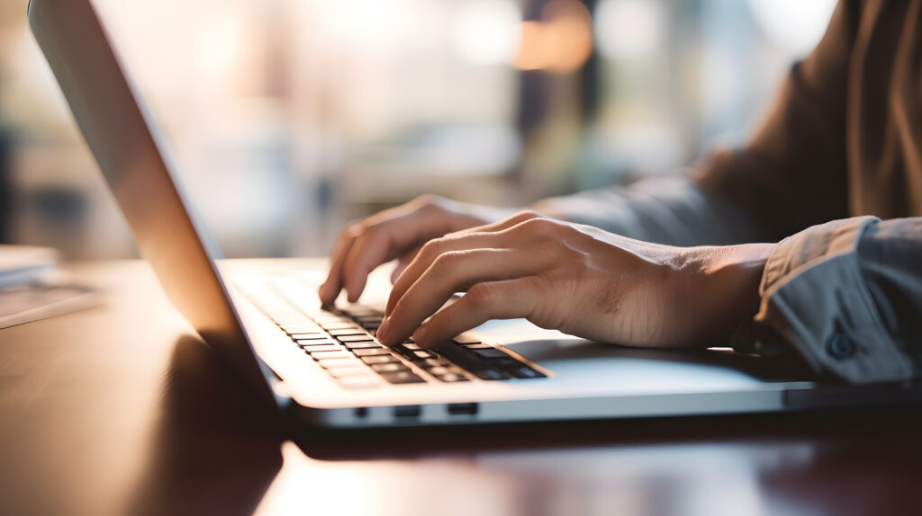Hands type on a laptop keyboard at a desk, illuminated by soft natural light coming from the background.