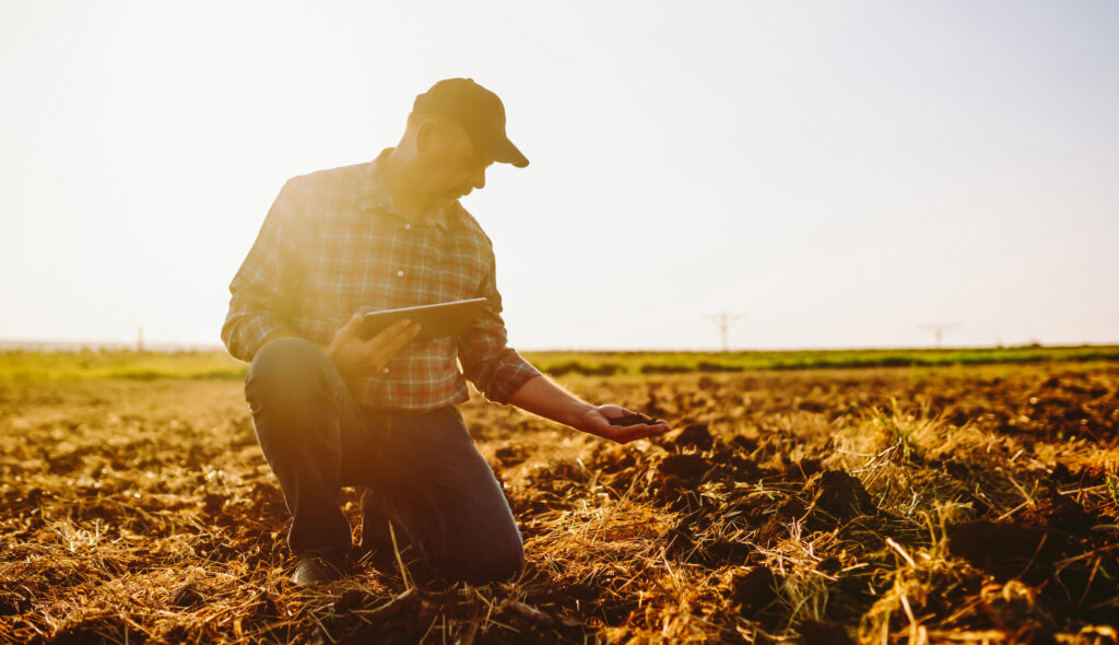 Kneeling farmer in a field holds soil in one hand, tablet in the other, sunlight shining behind.