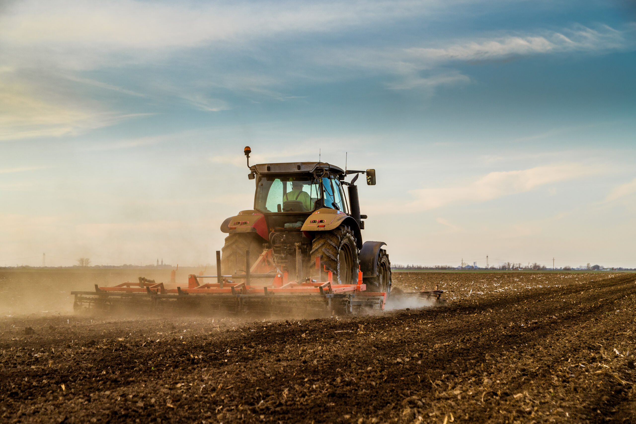 A tractor works across a broad, dusty field beneath a sky scattered with clouds.