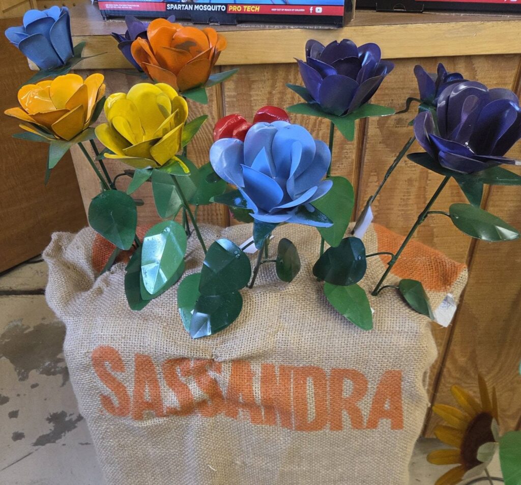 Colorful metal flowers arranged in a burlap sack labeled Sassandra, displayed on a wooden surface indoors.