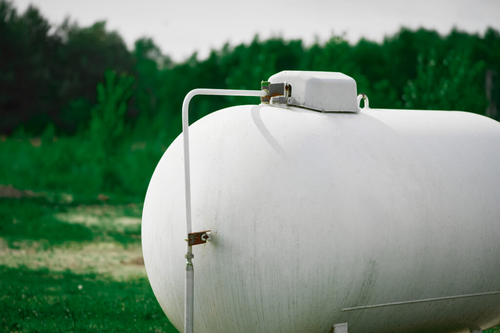 White propane tank sitting on green grass, surrounded by trees in the background under an open sky.