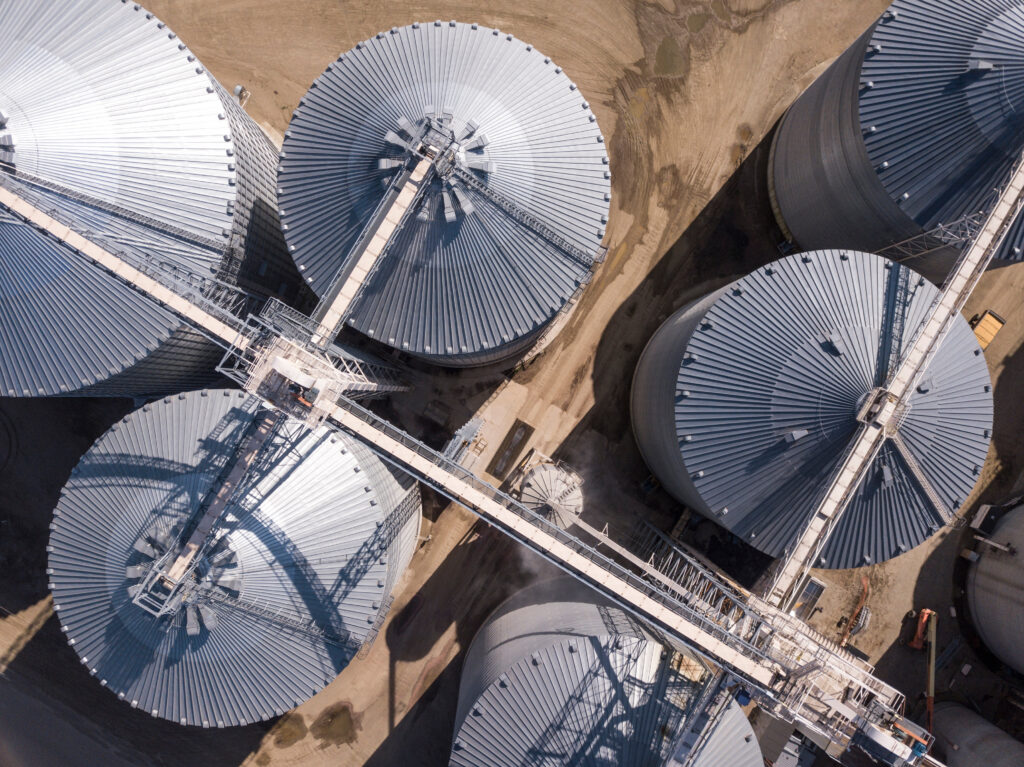 Aerial view of metal grain silos linked by walkways, set on a dirt surface.