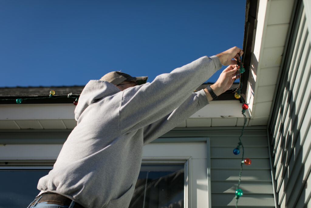 A person in a gray hoodie hangs colorful string lights along a house roofline on a bright, sunny day.