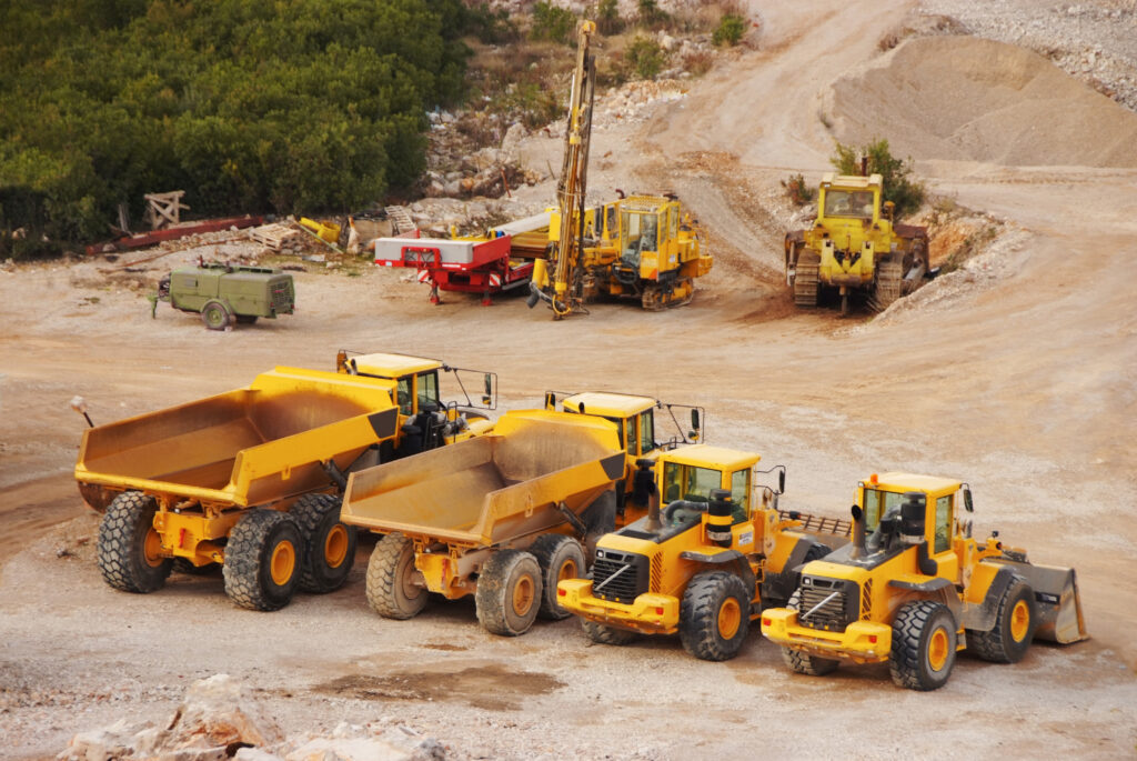 Yellow dump trucks, loaders, and excavation machines operate on a dirt construction site.