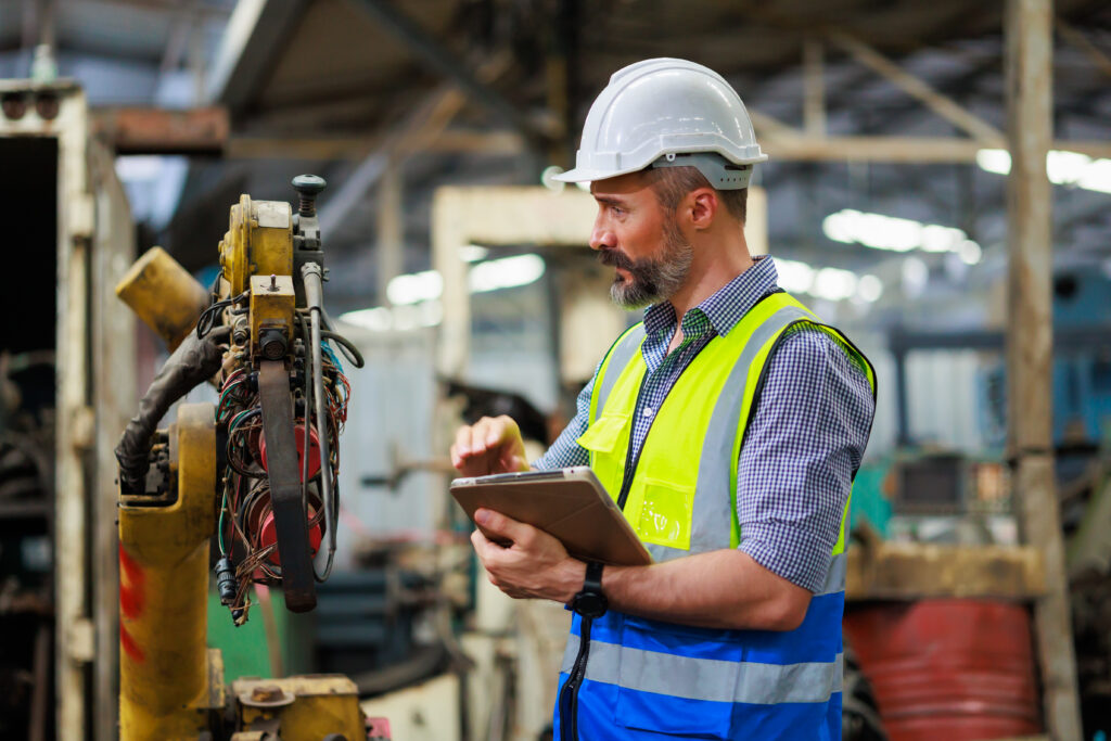 Man in hard hat and safety vest checks an industrial robot and uses a tablet inside a factory.