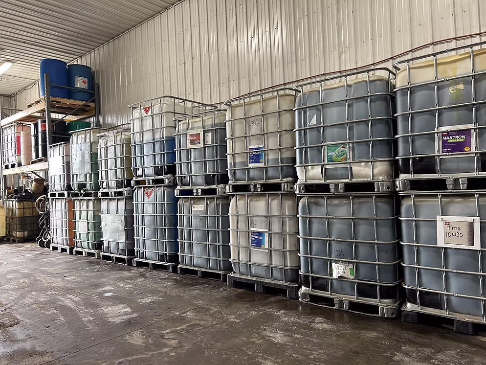 Plastic chemical containers stacked in a warehouse with metal walls and concrete floors.