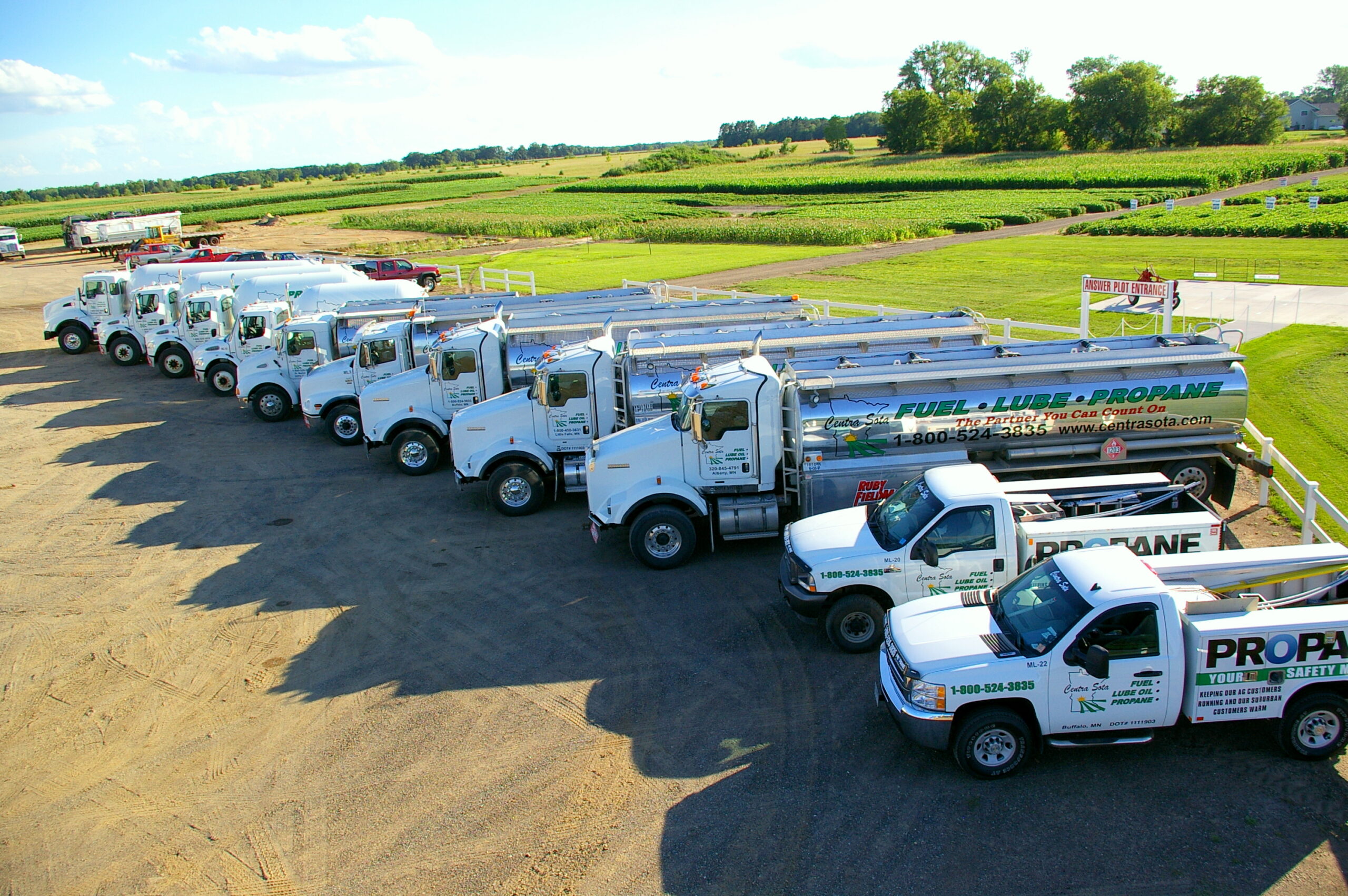 White propane trucks lined up on a lot beside green fields, all under a clear blue sky.