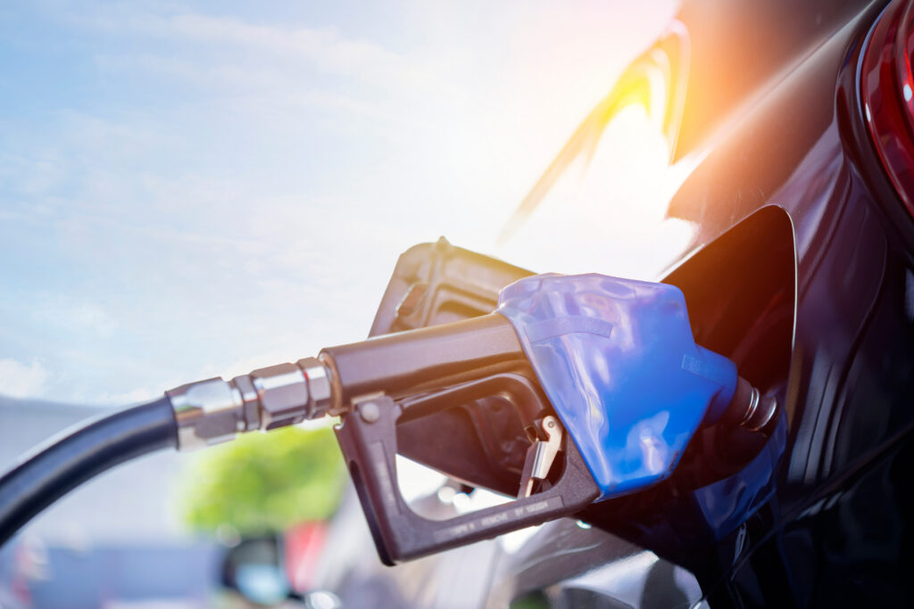 Blue fuel nozzle filling a car at a gas pump, sunlight shining in the background.