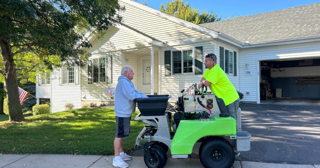 An older man speaks with a worker by a green machine outside a white house on a sunny day.