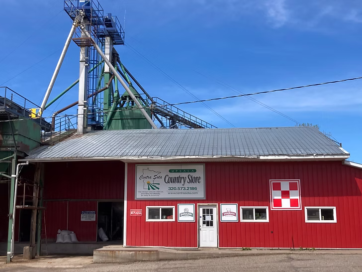 Red country store with metal roof, nearby industrial structures, and a clear blue sky behind them.