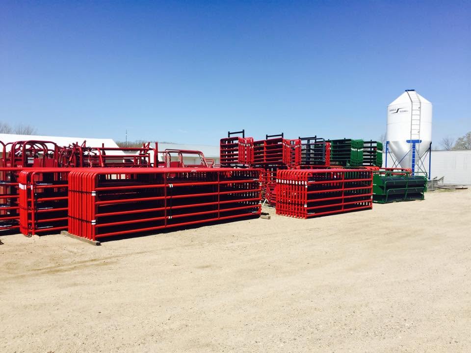 Red and green livestock panels stacked on gravel outside, with a white silo and blue sky behind them.