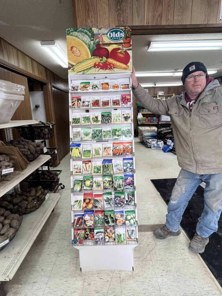 Man by seed packet display in a store, with vegetables and potatoes on shelves nearby.