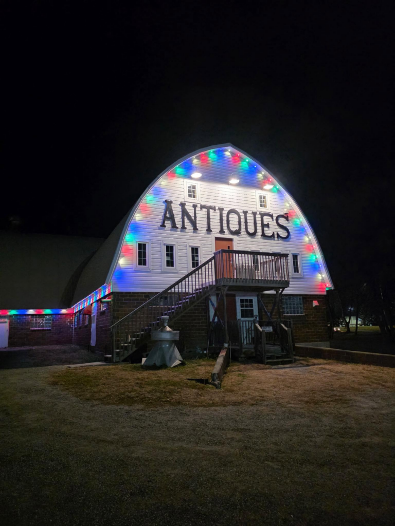 Barn-shaped antiques store at night, roof and entrance trimmed with colorful glowing lights.