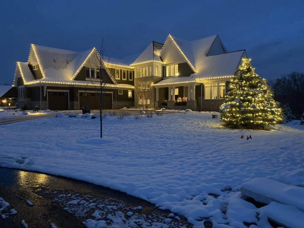 A snow-covered large house and tree glow with white Christmas lights on a winter evening.