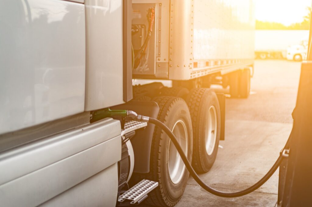 A semi-truck fuels up at a gas station while sunlight shines in the background.
