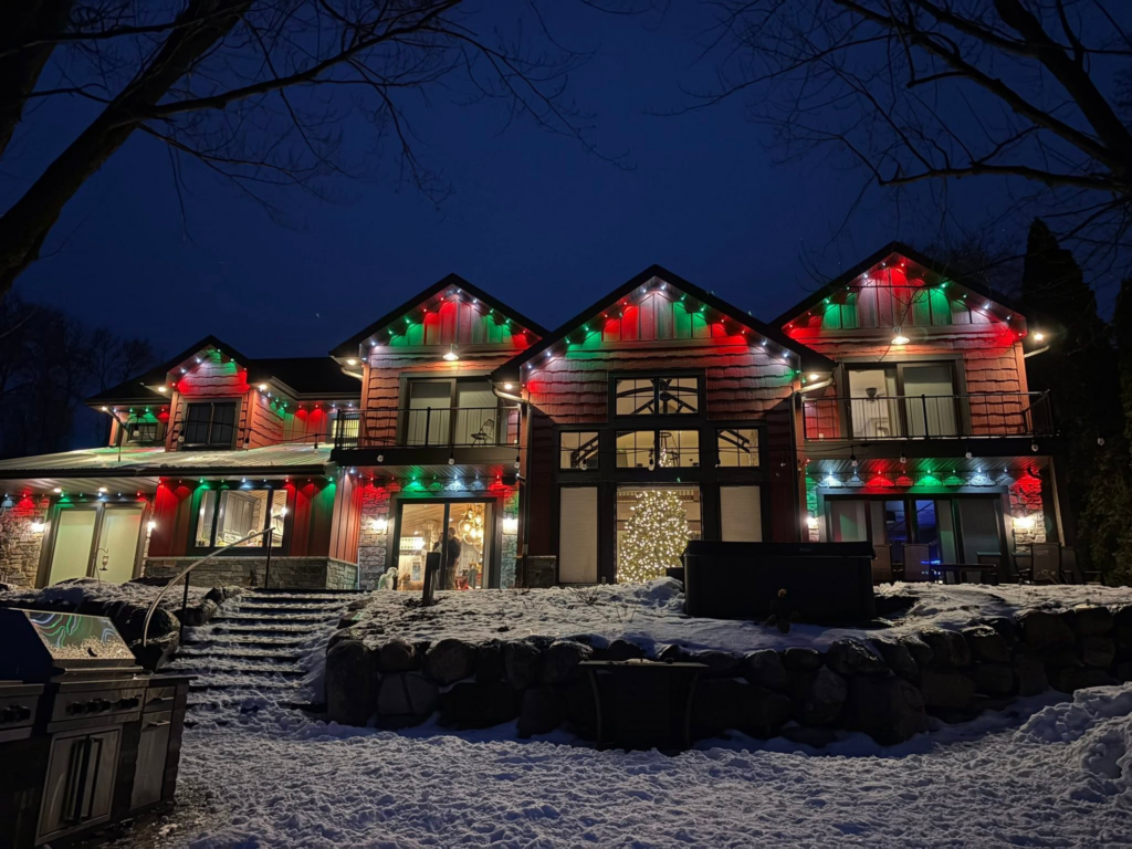 Snowy large house glowing at night with Christmas lights and a lit tree inside.
