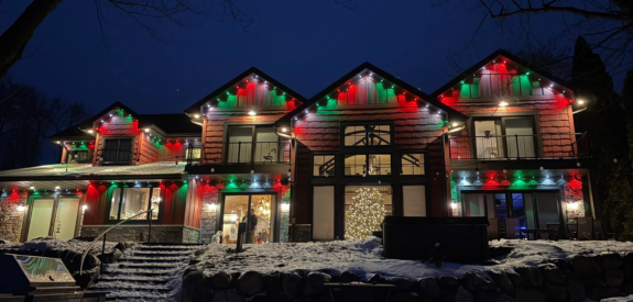 Snowy large house glowing at night with Christmas lights and a lit tree inside.
