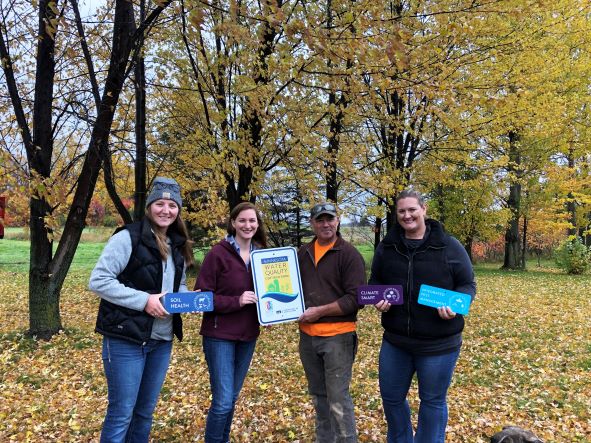 Four people outdoors in autumn hold soil health and climate signs, smiling together at the camera.