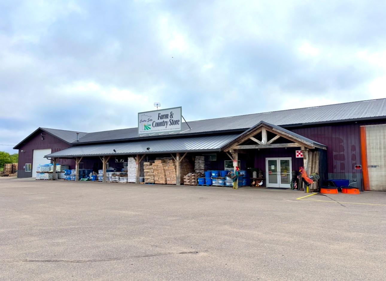 Farm and country store with porch, pallets, feed bags, and supplies visible outside on a cloudy day.
